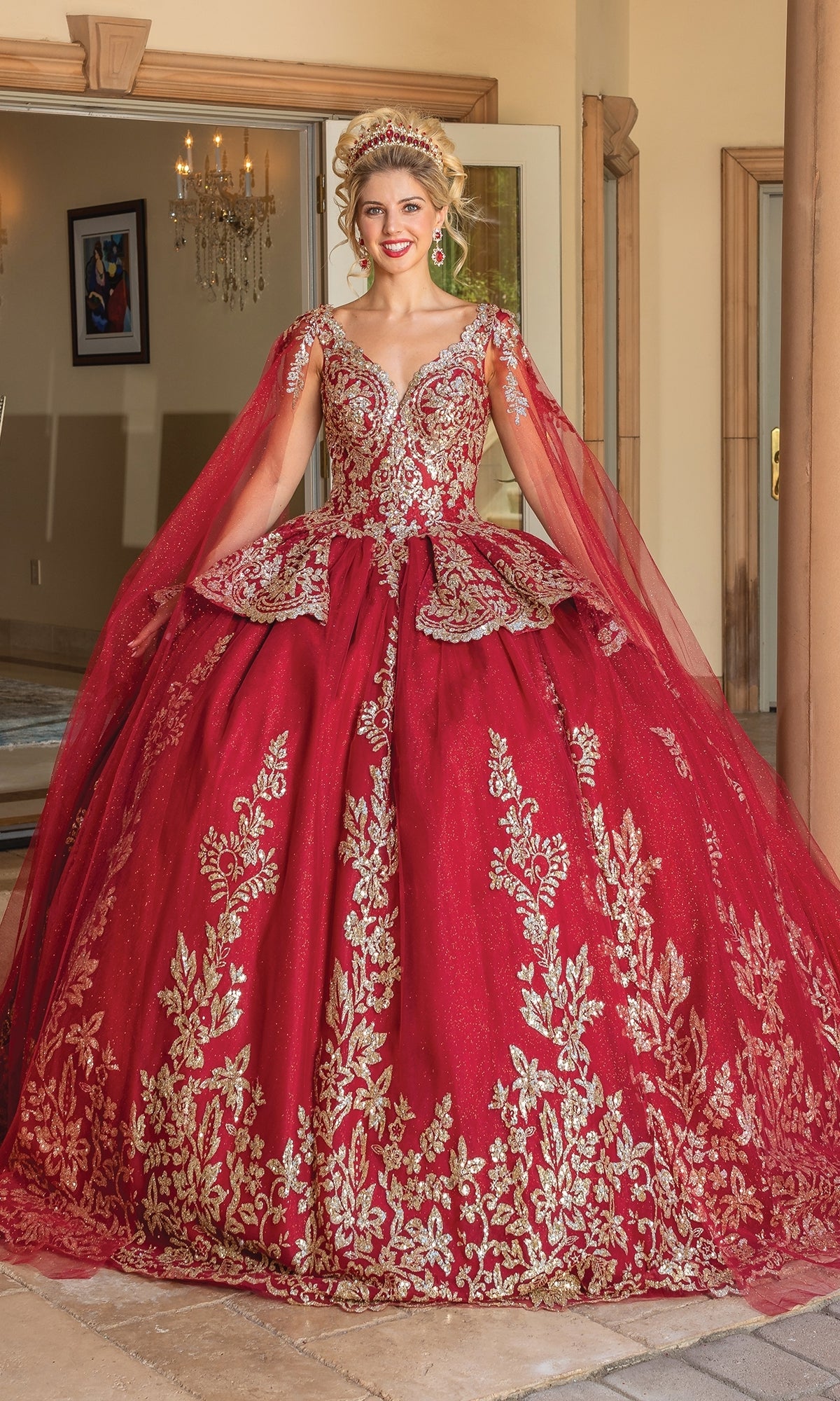 A woman smiles at the camera indoors wearing Dancing Queen's Quinceanera Dress 1781, a burgundy gown with gold embroidery, sheer cape sleeves, and a gold tiara.